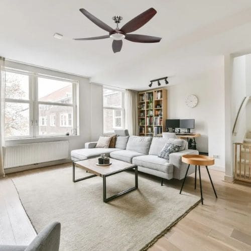 Salon lumineux avec ventilateur de plafond moderne, canapé gris, table basse et étagères, ambiance cosy et bien éclairée.